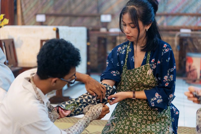 A candid photo of two participants during a batik workshop, preparing for the wax application process. The man is assisting the woman in putting on a protective sleeve typically used to prevent contact with hot wax. Both are wearing aprons with traditional batik patterns, seated indoors in a learning environment that highlights Indonesian cultural practices.