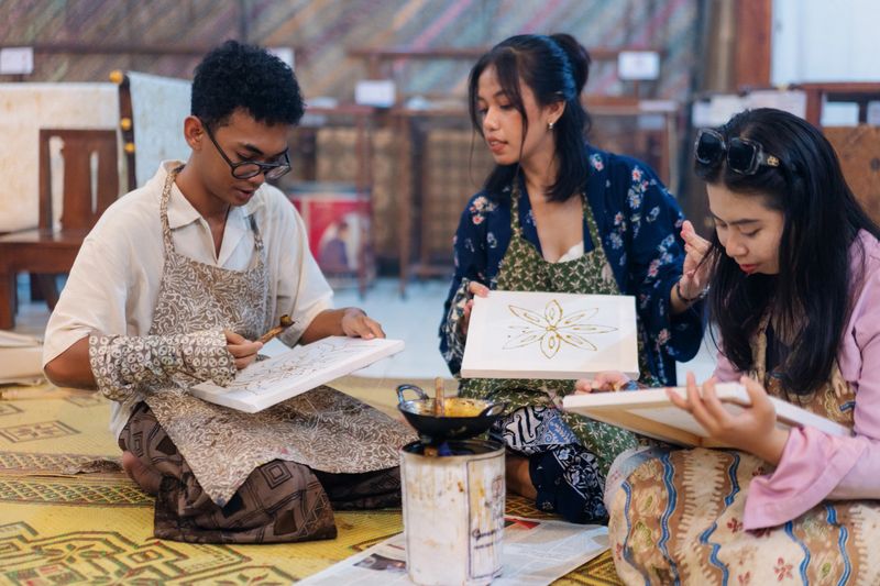A group of diverse friends participate in a traditional batik workshop in Indonesia, drawing intricate wax patterns on fabric using a canting tool and hot wax. Seated on woven mats, they share space and conversation while honoring a centuries-old textile tradition rooted in cultural heritage and slow craftsmanship.