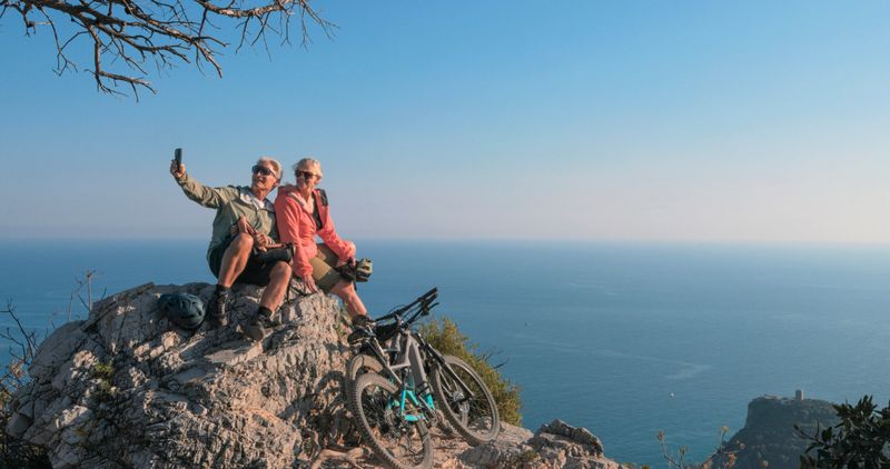 Mature couple with e-bikes take selfie on top of coastal viewpoint, Mediterranean Sea below, Liguria
