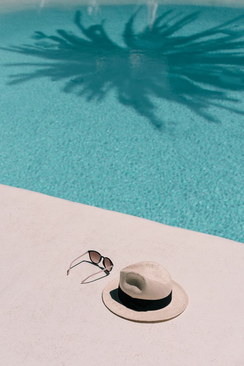 Straw hat, sunglasses at poolside. Summer vacation holidays. Swimming pool with a palm tree shadow