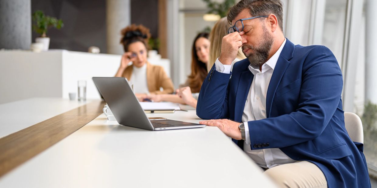A stressed man in a blue blazer rubbing his eyes at a conference table with a laptop.
