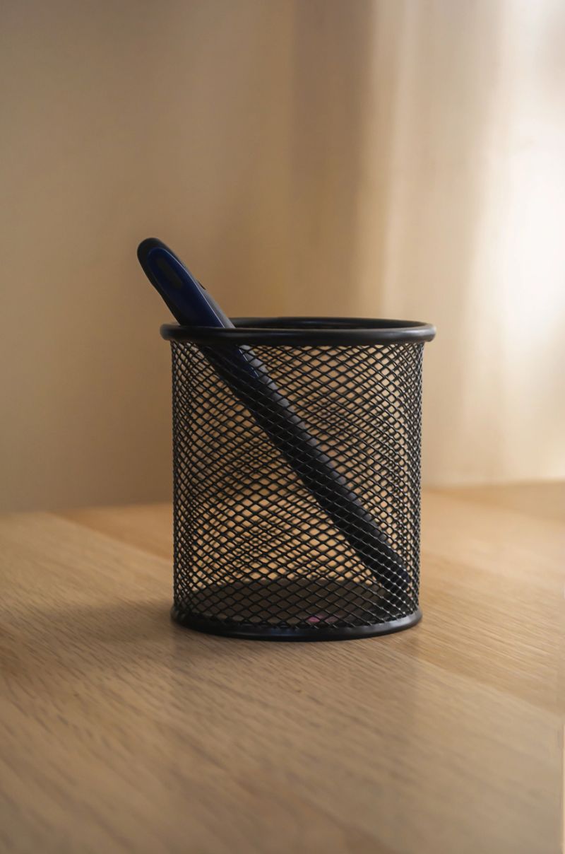 A minimalistic photo of a single black pen inside a black mesh metal pen holder placed on a wooden desk. Soft natural light illuminates the scene, creating a warm and cozy atmosphere with a neutral beige background
