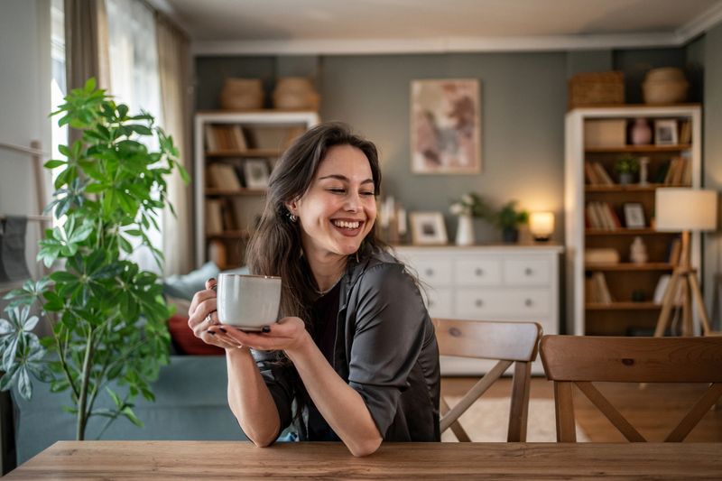 Young woman sitting at a dining table, holding a mug and smiling, savoring a peaceful moment of relaxation in a cozy living room filled with warm decor and natural light