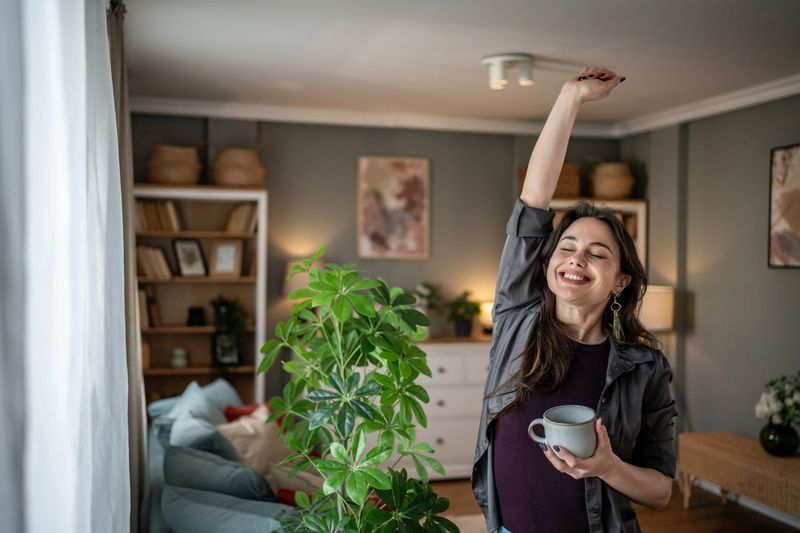 Young woman holding a coffee mug, stretching in her cozy living room, savoring a peaceful morning at home. Enjoying the tranquility of her weekend, surrounded by natural light and comfort