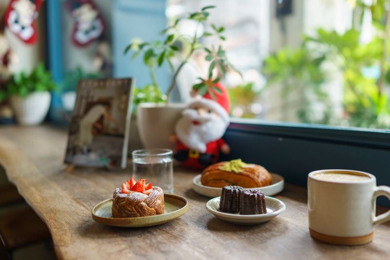 A festive cafe scene with a strawberry Danish, Cannelé, croissant, and coffee set on a wooden table near a window decorated with holiday decor and natural plants.