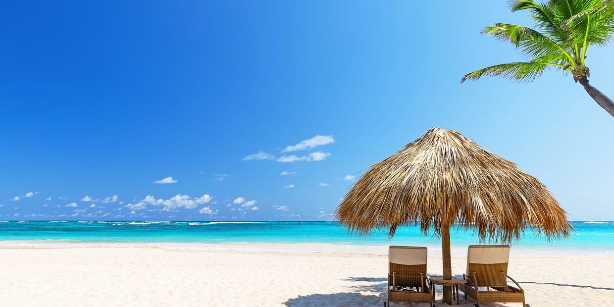 Two chairs under a straw umbrella on a sunny beach with clear blue water.