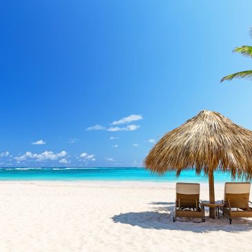 Two lounge chairs under a thatched umbrella on a sunny beach.