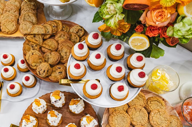 This vibrant dessert buffet is a feast for the senses, featuring an array of delectable treats. Fluffy cupcakes topped with white frosting, fresh raspberries, and dried orange slices sit elegantly on tiered stands. Freshly baked chocolate chip cookies are artfully arranged on wooden trays, adding a rustic charm. Glass jars brim with colorful candies, complementing the citrus theme. A bouquet of roses and greenery, accented with lemon and orange slices, serves as a stunning centerpiece. The spread, draped with a crisp white tablecloth, exudes a festive and inviting atmosphere, perfect for any celebration.#Pastries #Indulgence #Gourmet #Foodie #Delicious #Yummy #Tasty #Snack #Event #Gathering #Catering #Decor #Elegant #Colorful #WhiteFrosting #WoodenTrays #GlassJars #Sweets #Bakery #Homemade #Festive #Treats