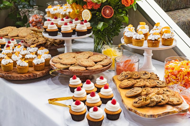 This vibrant dessert buffet is a feast for the senses, featuring an array of delectable treats. Fluffy cupcakes topped with white frosting, fresh raspberries, and dried orange slices sit elegantly on tiered stands. Freshly baked chocolate chip cookies are artfully arranged on wooden trays, adding a rustic charm. Glass jars brim with colorful candies, complementing the citrus theme. A bouquet of roses and greenery, accented with lemon and orange slices, serves as a stunning centerpiece. The spread, draped with a crisp white tablecloth, exudes a festive and inviting atmosphere, perfect for any celebration.#Pastries #Indulgence #Gourmet #Foodie #Delicious #Yummy #Tasty #Snack #Event #Gathering #Catering #Decor #Elegant #Colorful #WhiteFrosting #WoodenTrays #GlassJars #Sweets #Bakery #Homemade #Festive #Treats