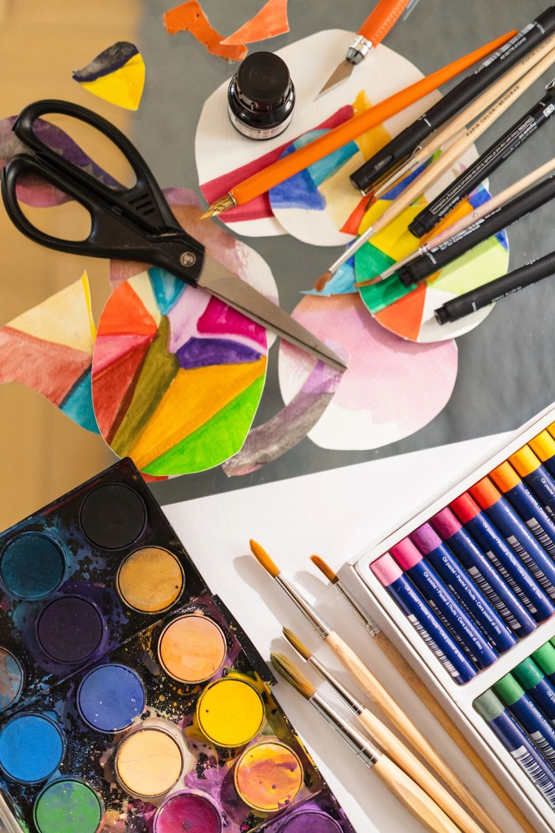 Top view of a vibrant and artistic setup featuring watercolor paints, oil pastels, brushes, scissors, colored paper cut-outs, ink, and fine liners. A perfect representation of a creative workspace, DIY projects, or school art supplies.