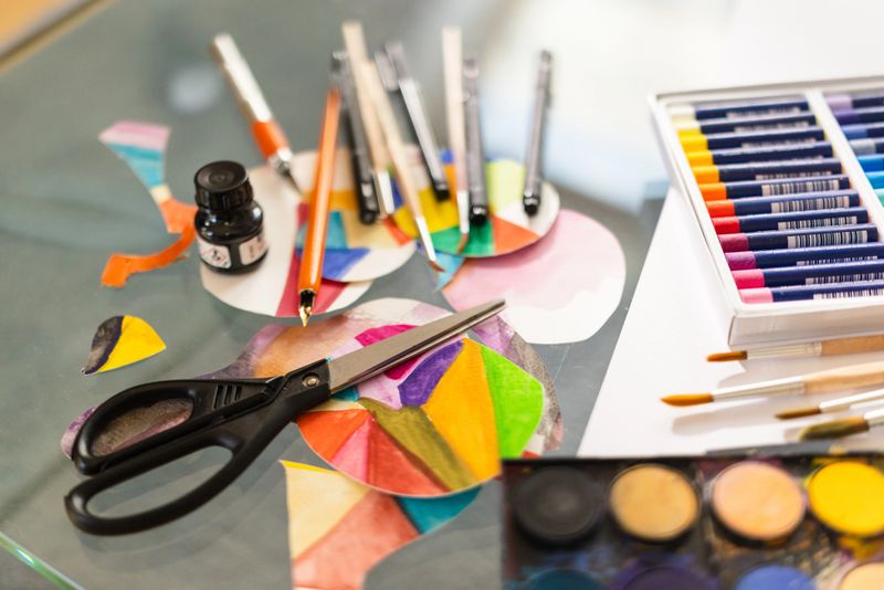 Close-up image of watercolor paint pans with vivid splashes, paintbrushes, and pastel sets in the background. Represents hands-on creativity, expression, and colorful experimentation.