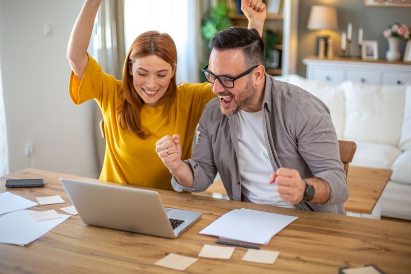 Happy couple sitting at home table, looking at laptop cheering, screaming, holding hands up, their favorite team is winning.