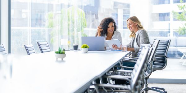 Two women collaborating over a tablet in a bright modern conference room.