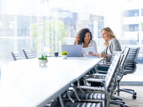 Two women collaborating over a tablet in a bright modern conference room.
