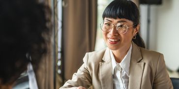 Woman in glasses and beige blazer smiling during a conversation.