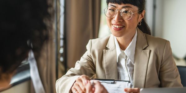 A professional woman smiles while handing a clipboard to another person.
