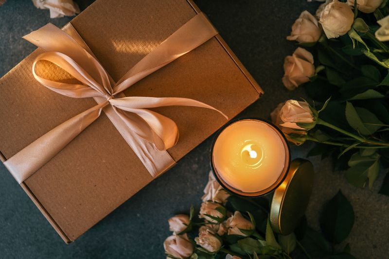Top view of a glowing candle, beige roses, and a gift box tied with satin ribbon, arranged on a dark stone background in a cozy setting.