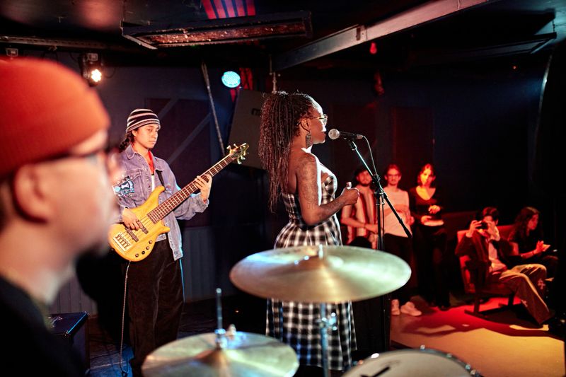 Focus on mature Black chanteuse in plaid strapless dress at microphone, female guitarist to her left, percussionist in foreground, partial view of audience.