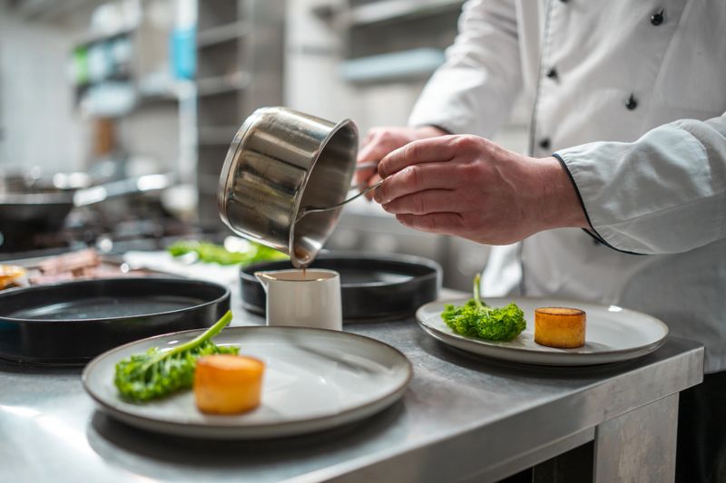 A professional chef carefully plates gourmet dishes in a bright, modern kitchen. The scene features precise hand movements, vibrant green vegetables, and minimalistic plating on white ceramic plates under soft, even lighting.