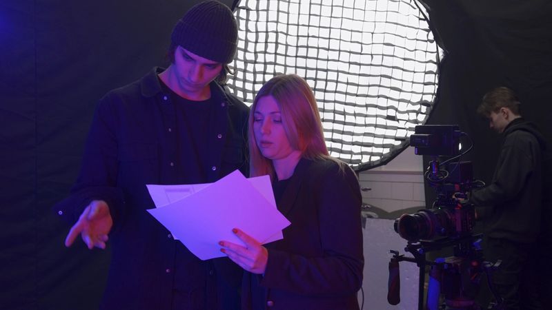 A female producer and videographer discuss the storyboard in the studio before filming. Meanwhile, the lighting technician adjusts the lighting equipment. Behind the scenes of filming video production.