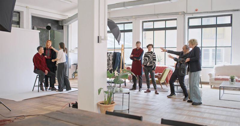 Wide-angle view of diverse production team working in spacious industrial studio. Young professionals in casual attire coordinate lighting and positioning while models prepare for photoshoot.