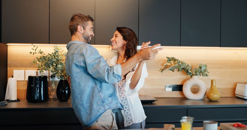 Mature couple, smile and dancing in kitchen with love, bonding and celebration for anniversary in home. People, man and woman with holding hands, excited or playful rhythm for relationship at house