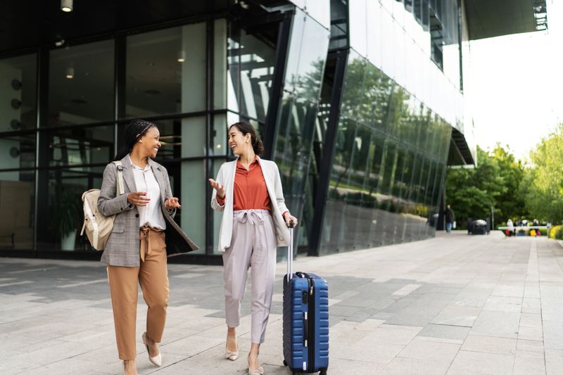 Two cheerful businesswomen are walking and talking outside a modern office building, one pulling a suitcase, discussing work and travel plans