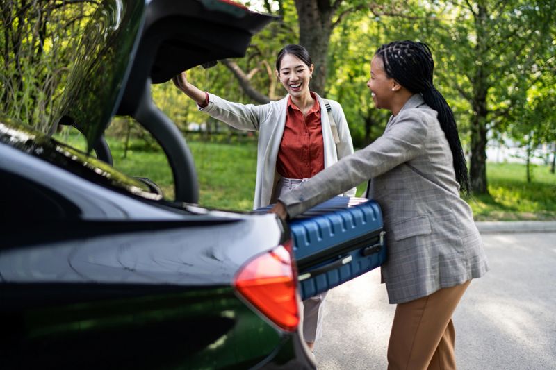 Two cheerful businesswomen loading a suitcase into the trunk of a car, preparing for an exciting business trip, sharing smiles and enjoying the moment together in a vibrant parking lot