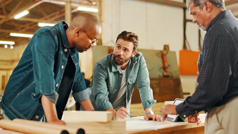 Carpentry, talking and men in workshop planning for furniture, wood restoration and lumber manufacturing. Construction, factory and carpenters in meeting with planks, timber and board for renovation
