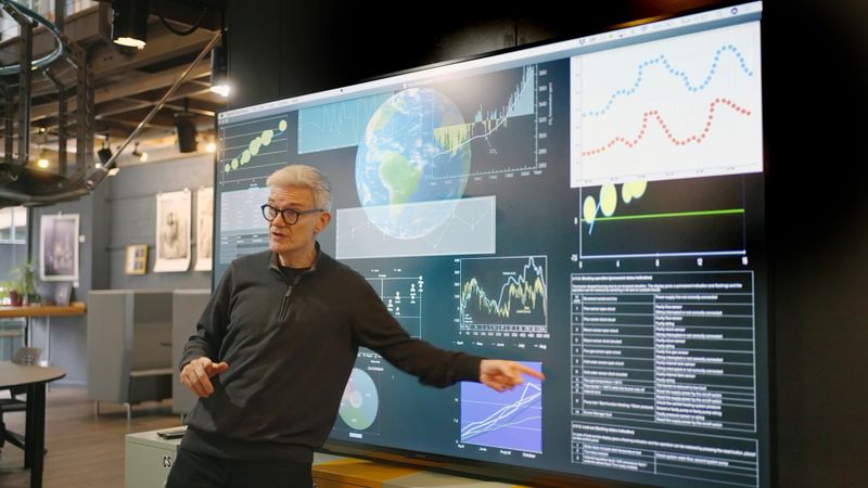 Stock photograph of a mature man giving a lecture in front of a large screen depicting data associated with the planet earth, ie climate change etc .