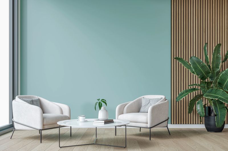 This image showcases a modern, minimalist seating area with two elegant white armchairs positioned around a sleek, low-profile white coffee table in front of a celeste (light) blue wall background. A wooden slat accent wall on the right adds warmth and texture, complemented by a large green plant in a black pot. Natural light from the floor-to-ceiling window enhances the soft, neutral tones, creating a bright and inviting atmosphere. 3D rendered image.