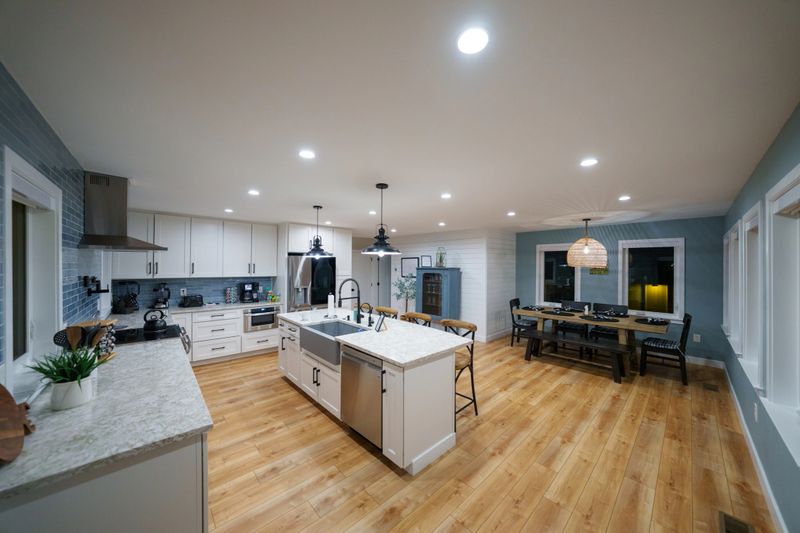 Wide shot of a modern farmhouse style dining area and kitchen with stainless steel appliances, kitchen island, white cabinetry, and blue tile backsplash in a middle-class home in Maryland, USA.