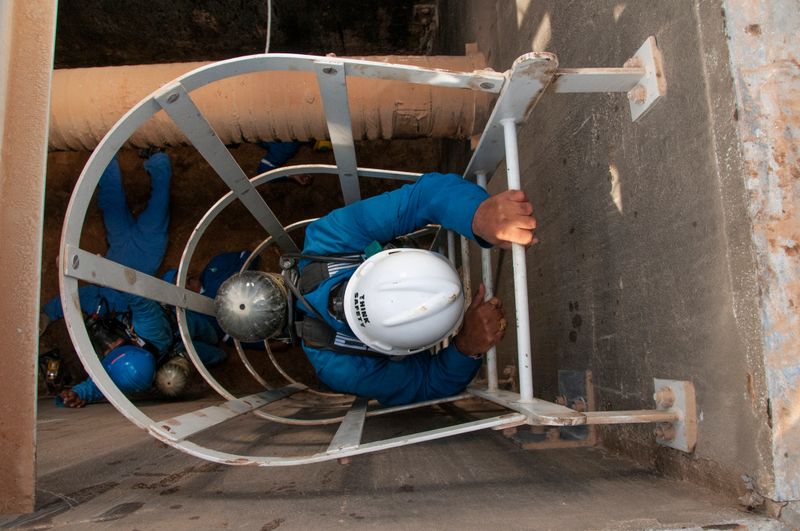 Workers on a rescue mission wearing safety gear, including a hard industrial helmet written Think Safety and breathing apparatus, descend a vertical metal ladder into a confined industrial space, emphasizing safety procedures in hazardous environments
