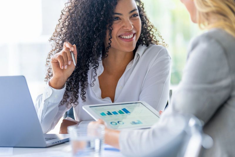 Two business women meeting and looking at financial data, charts and graphs on a digital tablet and a laptop computer. One woman is happy and smiling and pointing with a pen