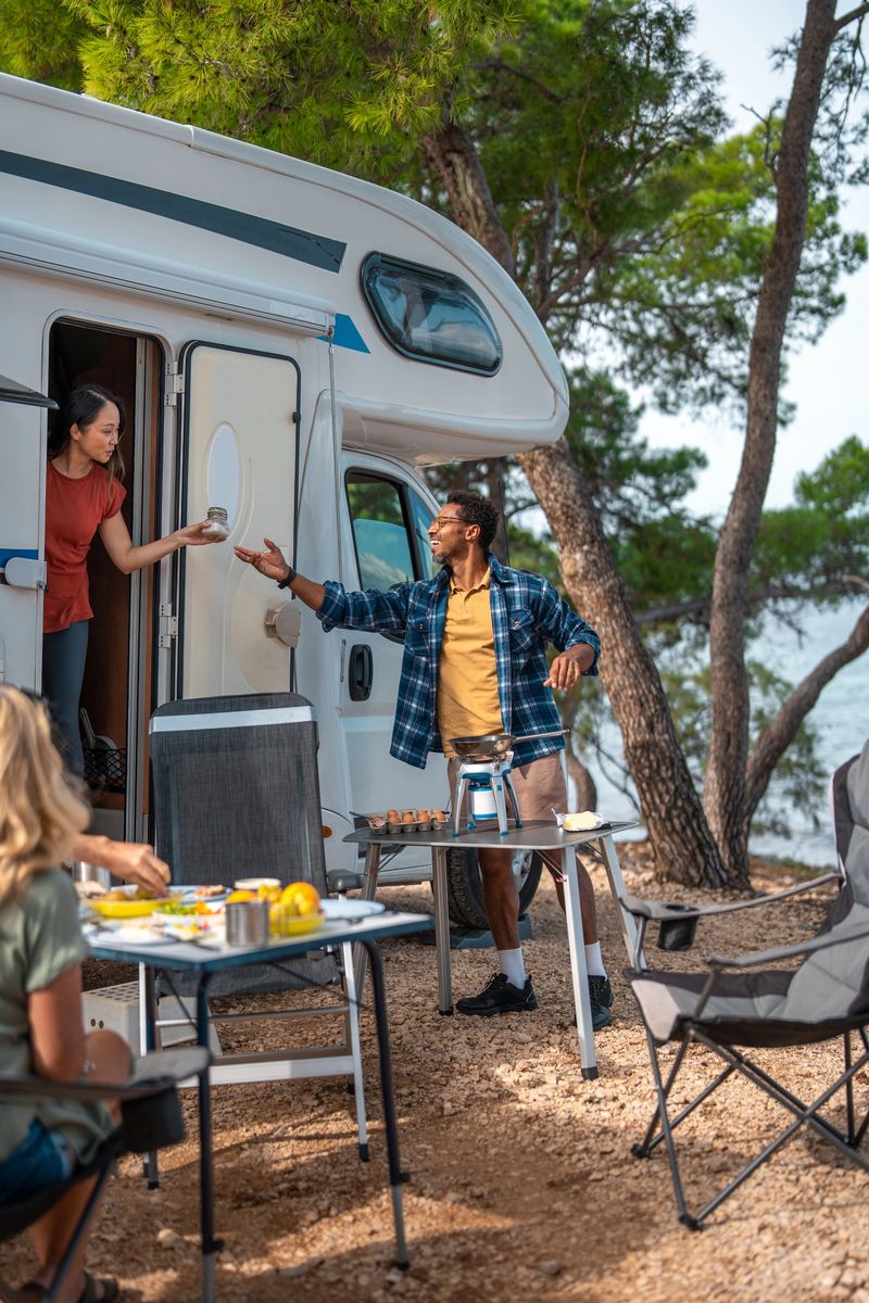 Black man in casual attire prepares breakfast at a seaside campsite, using portable camping table outside motorhome, creating an authentic outdoor living experience.