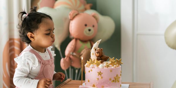 A toddler blowing out candle on a pink teddy bear-themed birthday cake.