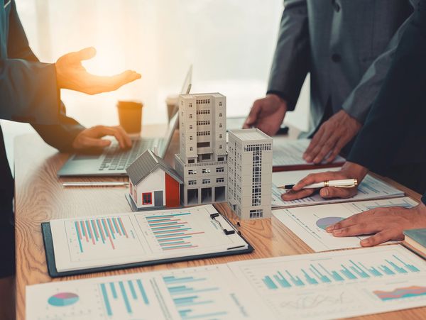People discussing real estate investment with building models and charts on a desk.