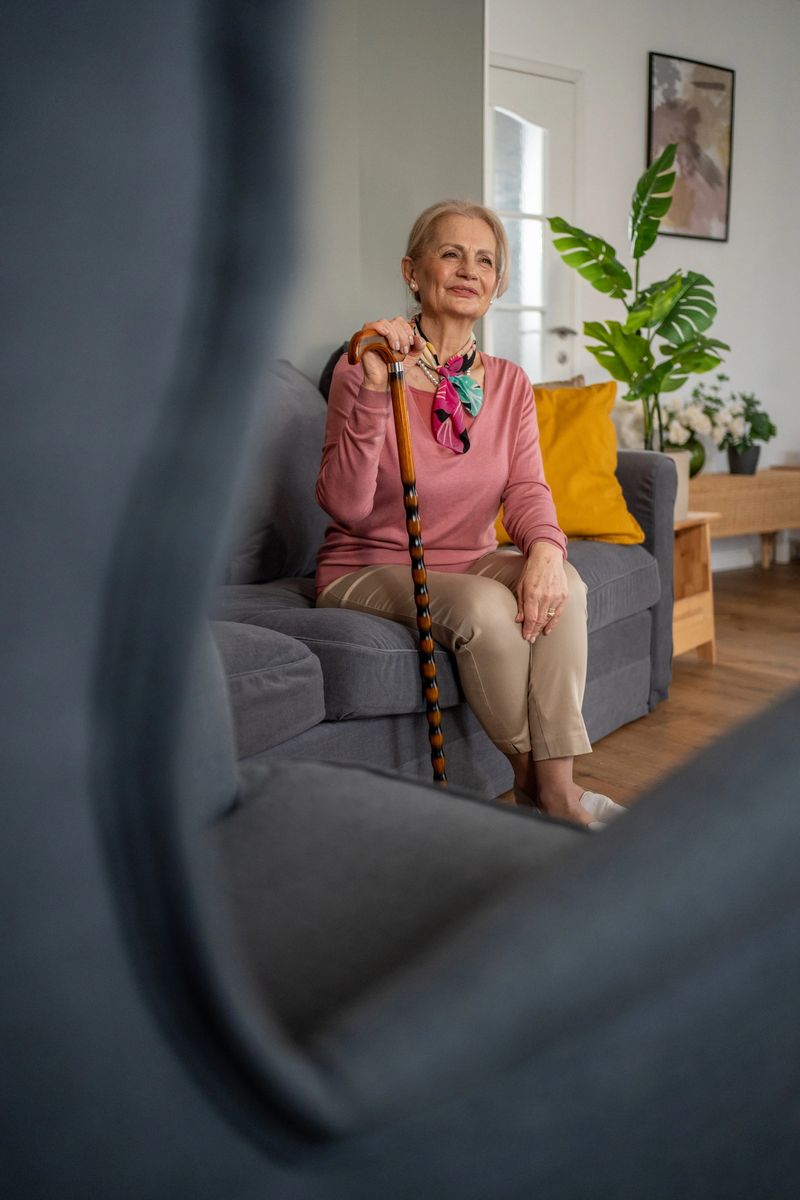 Happy senior woman sitting comfortably on a sofa in her living room, holding a walking cane and gazing thoughtfully into the distance, embodying tranquility and contentment