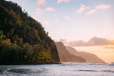 Sunset over lush cliffs by the ocean with gentle waves.