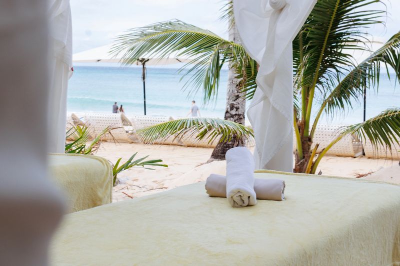 A tranquil beachfront massage setup features two massage tables under a canopy, surrounded by palm trees and the ocean in Boracay. Soft linens sway in the ocean breeze, creating a serene escape.