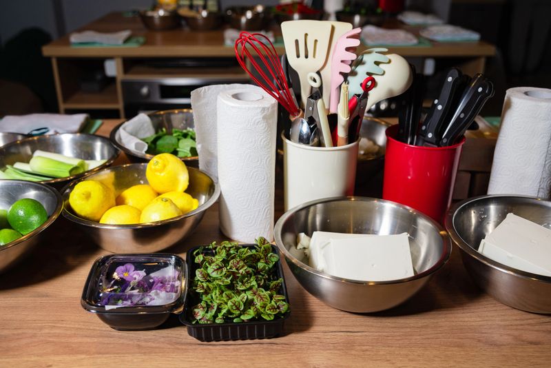 Fresh tofu, edible flowers, microgreens, and lemons arranged on a cooking prep station with utensils and bowls.
