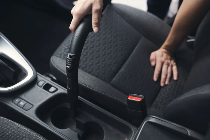 Person using a vacuum cleaner to clean the interior of a car, focusing on seat and central console areas. Depicts detailed cleaning and maintenance of the vehicle for hygiene and cleanliness.