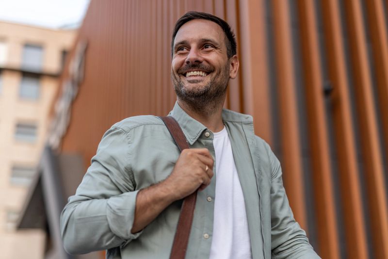 Cheerful businessman walking confidently near modern office buildings, enjoying a sunny day while glancing away, casually holding a shoulder bag and radiating positivity