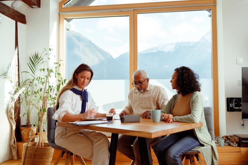 A woman, likely a financial advisor, is seated with a senior couple in a bright room with a stunning mountain view, indicative of a home in Slovenia. They are engaged in conversation, with documents laid out on the table, pointing to a discussion about pension insurance. The advisor is presenting information, while the couple listens intently, considering the options for their retirement planning.