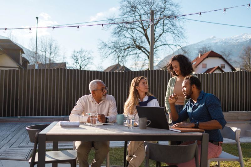 A family is gathered around a laptop outdoors, engaged in a serious business discussion, with a backdrop of mountains suggesting a balance between work and nature.