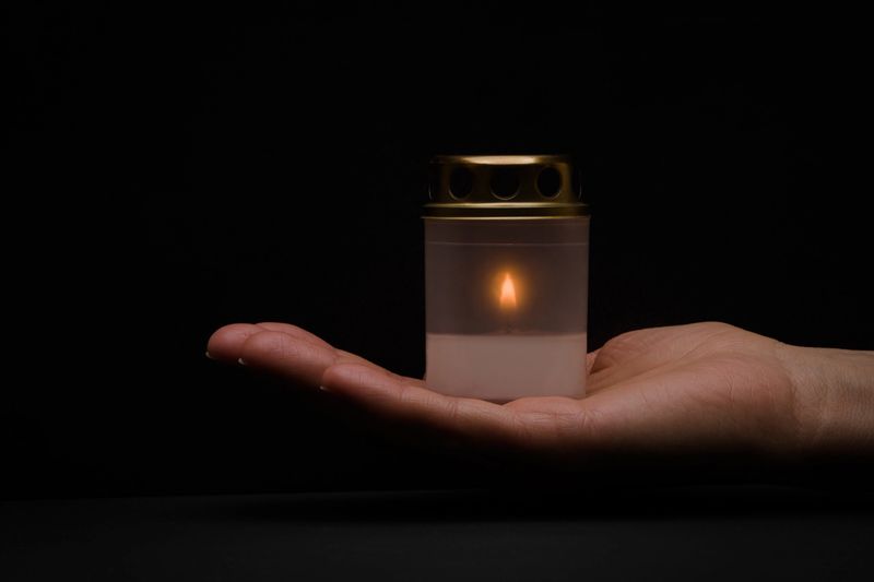Adult woman opened palm holding plastic container with white burning candle on dark black background. Closeup. Side view.
