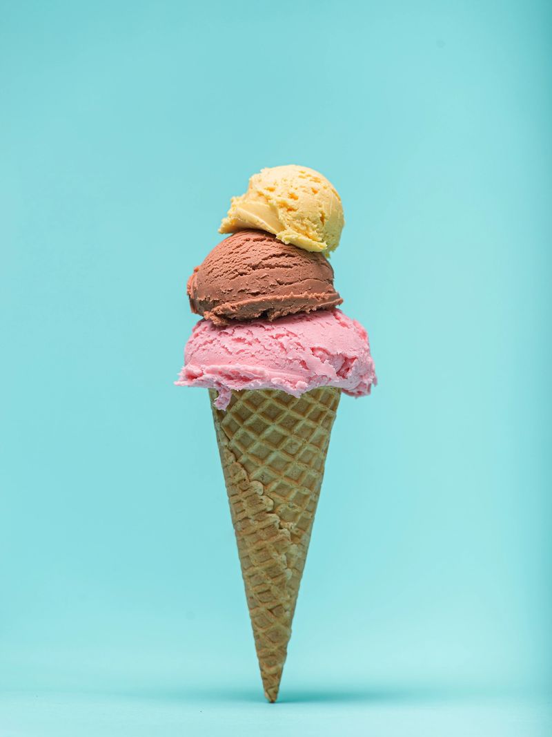A waffle cone overflows with three scoops of ice cream: strawberry, chocolate, and vanilla. The vibrant colors pop against a pastel blue background