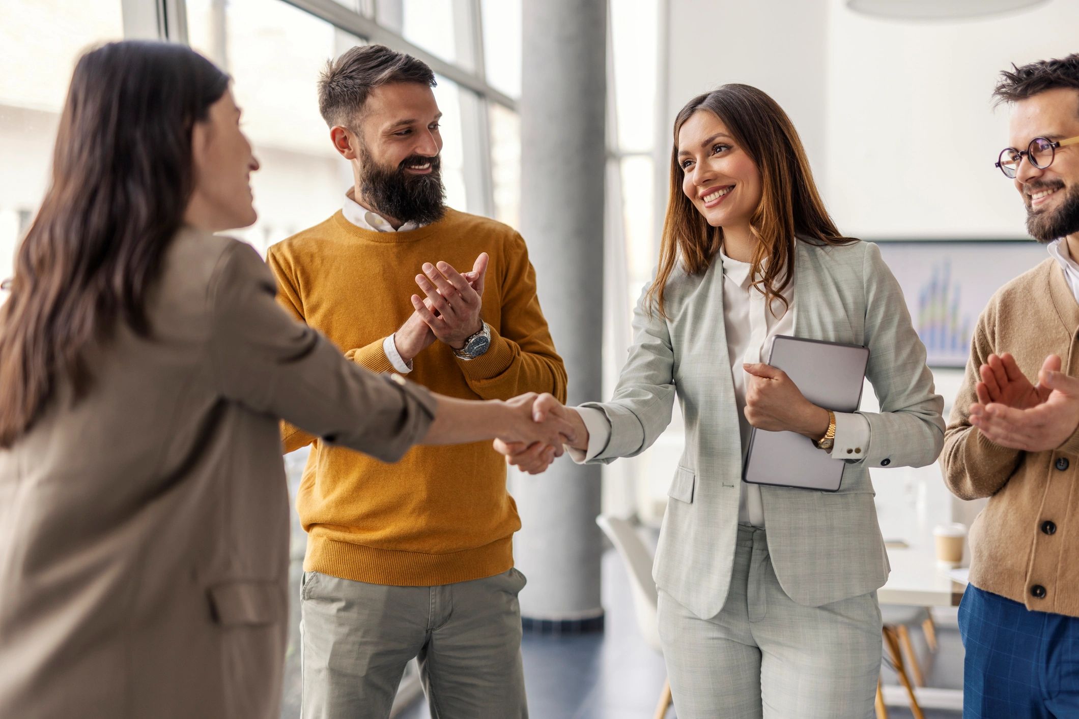 Two women shaking hands while colleagues applaud in a modern office.