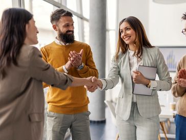 Two women shaking hands while colleagues applaud in a modern office.