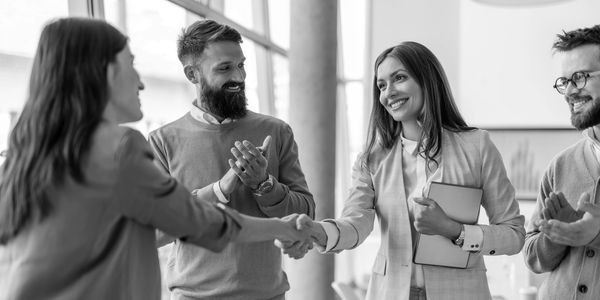 Two women shaking hands in a business setting with colleagues applauding.